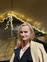 A picture of Mily. She has blonde hair that has a faded pink effect. Her hair is shoulder length. She is wearing a light cream jacket with a dark fleece underneath. The background is a marquee structure with fairy lights.