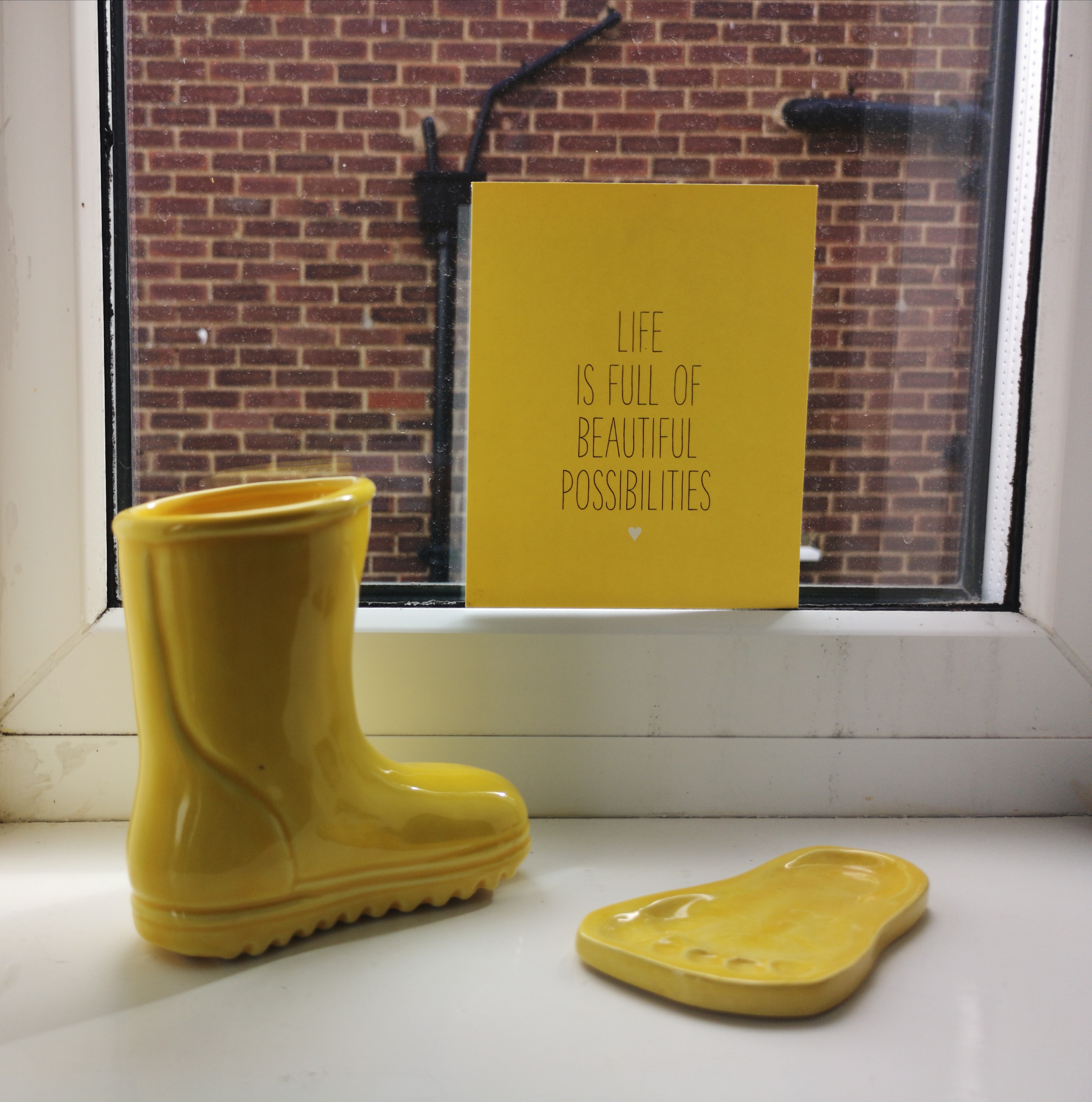 Yellow themed objects placed on a windowsill. Yellow Ceramic boot and footprint. And a yellow poster reading 'Life is full of beautiful possibilities.