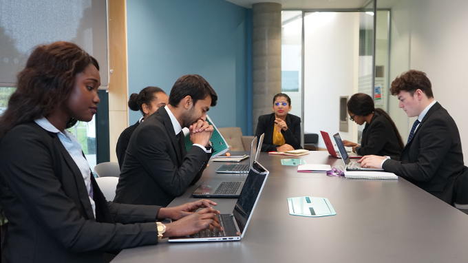 students sit around a table. They are smartly dressed and some work on their laptops