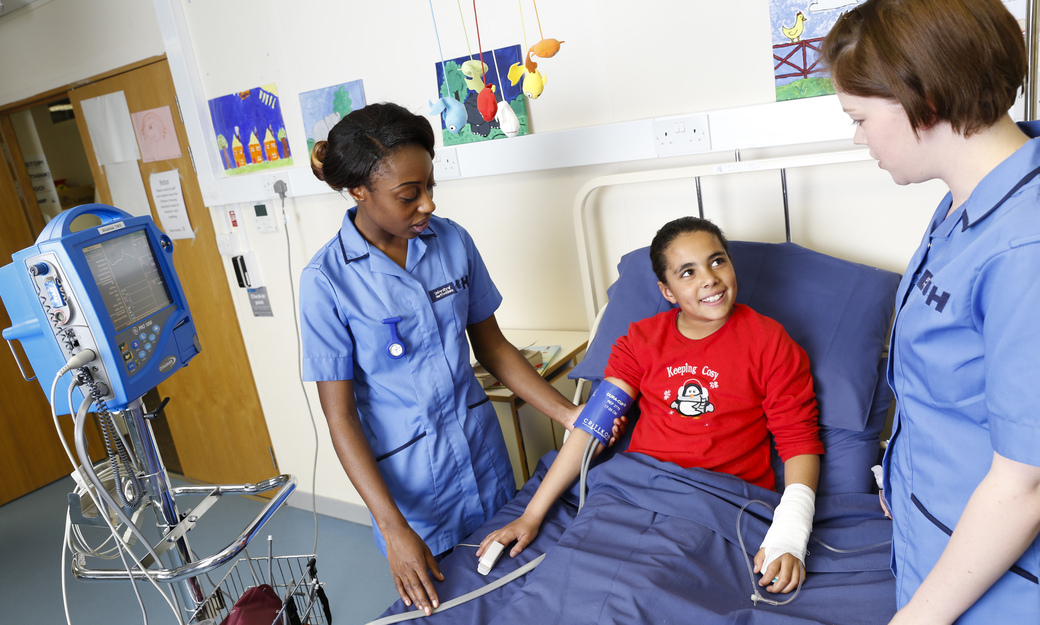 two student nurses taking care of a child in a ward bed and taking her blood pressure readings