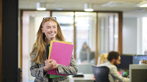 Female employee smiling whilst holding a collection of coloured files in an office.