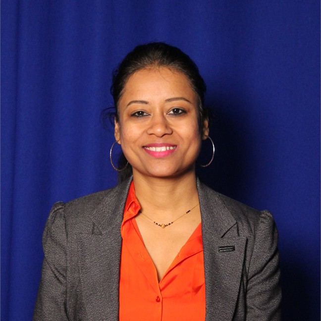 Shanti has dark hair, tied up in a ponytail, with gold hoop earrings, pink lipstick and a grey blazer over the top of an orange shirt, against a blue background
