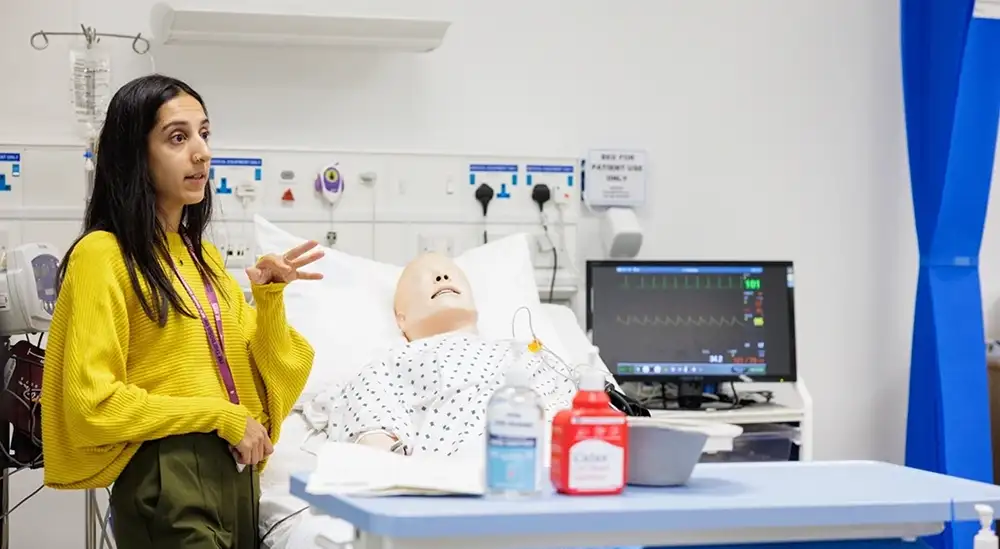 A lecturer talking while standing next to a hospital bed with a dummy in it