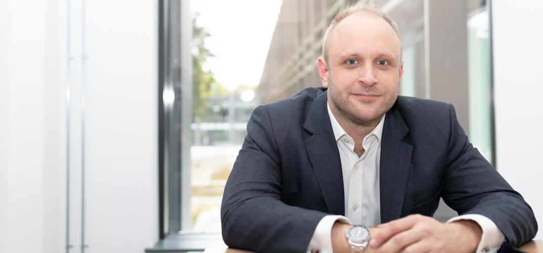 Professor Tim Vorley OBE sits at a desk in front of a window, wearing a suit and smiling