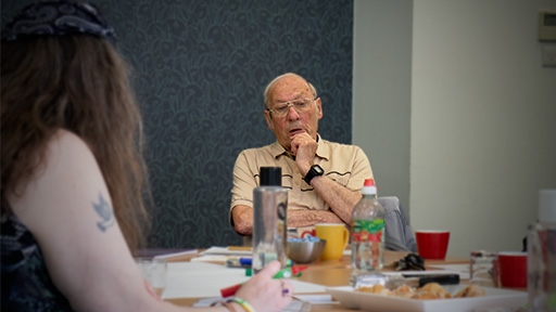 An older man sits with his hand on his chin, behind a table.  