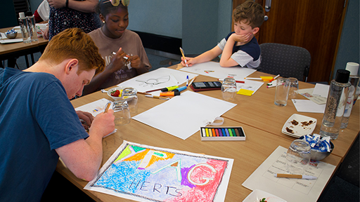 A group of three young people work on a table. Designing a logo which reads YPAG Herts with bright colours