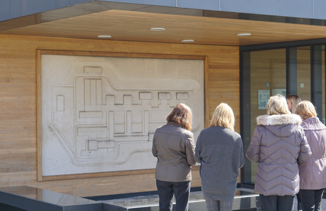 A group of women stand and look at 'College Plan' by Trevor Tennant, a wall relief sculpture carved out of white stone depicting the original geometric layout of the Main Building.