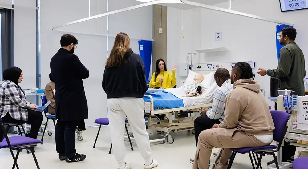 A lecturer talking to a group while standing next to a hospital bed with a dummy in it 