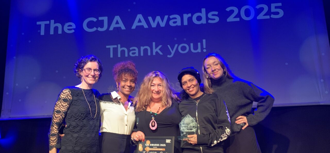 A group of five people standing together on stage at an awards ceremony, holding a certificate and a glass trophy. Behind them is a large screen displaying the text “The CJA Awards 2025 Thank you!” in bold white letters on a blue background. The individuals are dressed in a mix of formal and smart-casual attire, and the setting is brightly lit, indicating a celebratory event.