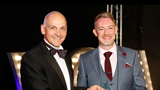 A man wearing a grey suit and a red tie smiles as he receives his award from Quintin McKellar