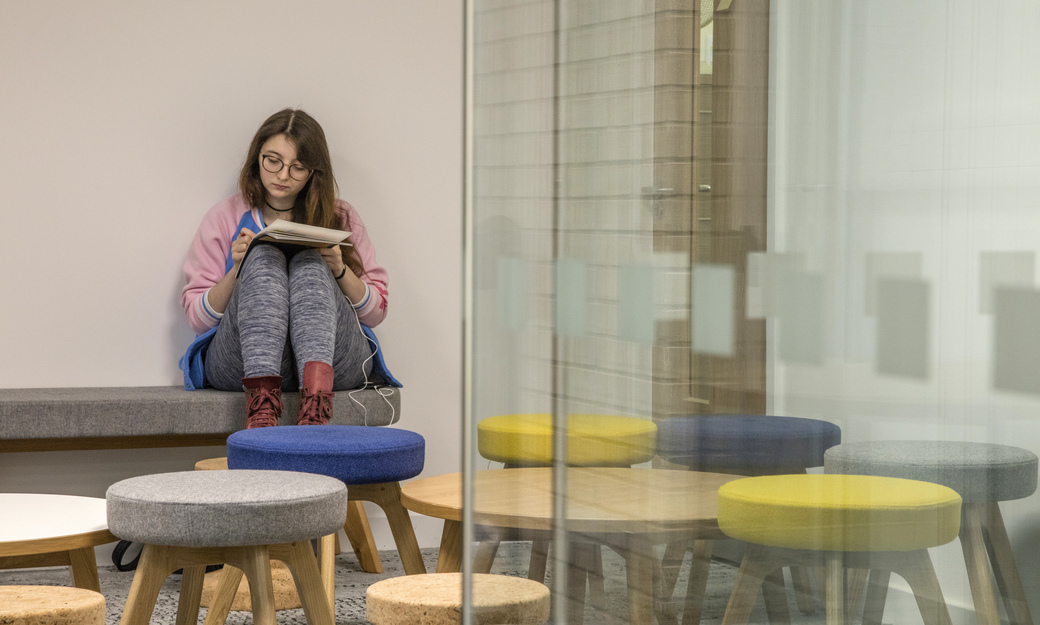 a female student making notes on a fabric covered bench, in a classroom with comfy stools and tables.