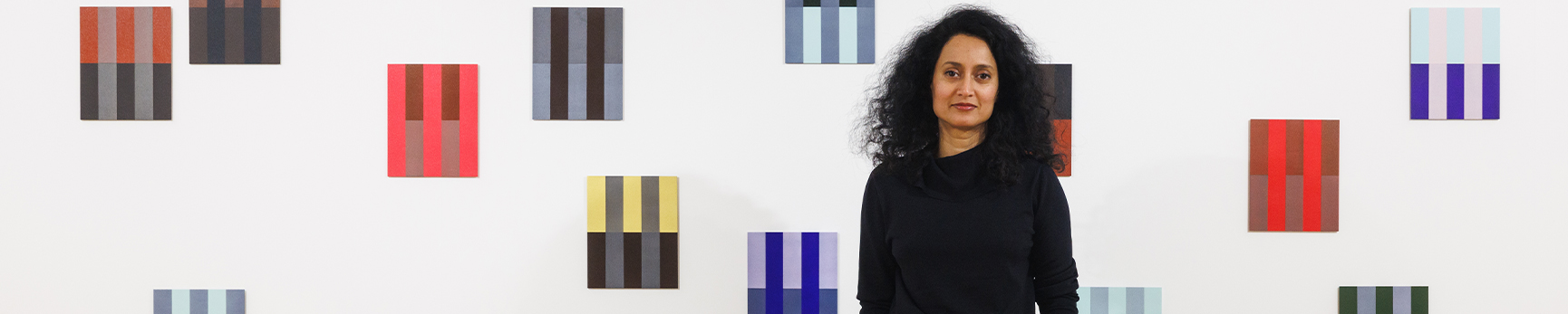 Rana stands in front of her 20 small rectangle geometric panel paintings against a white wall.