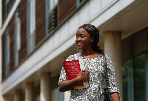 Very smiley Herts postgraduate student walking on campus and holding a text book in her arm