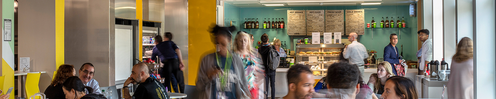 Photograph of the busy Cafe Rore on the College Lane Campus, filled with students, staff and visitors on the tables eating and drinking. Staff in the background are serving food and drink at the counter..