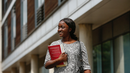 Very smiley Herts postgraduate student walking on campus and holding a text book in her arm