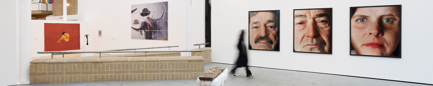 White gallery space with three large close up portraits and an abstract sculpture in the foreground, another large photograph and painting can be seen further away