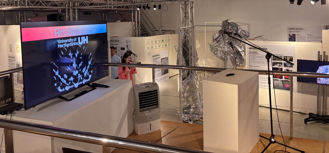 Exhibition space featuring University of Hertfordshire’s Art + Science Lab display on a large screen, surrounded by informational panels, sculptural installations, and technical equipment.”