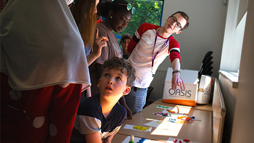 A tall brown haired man talks to a group of children who are looking at pieces of art on a table. 
