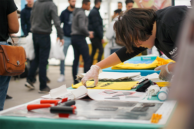 A volunteer with short brown hair and a black T shirt assisting with lino printing patterns onto a yellow UH Arts tote bag.