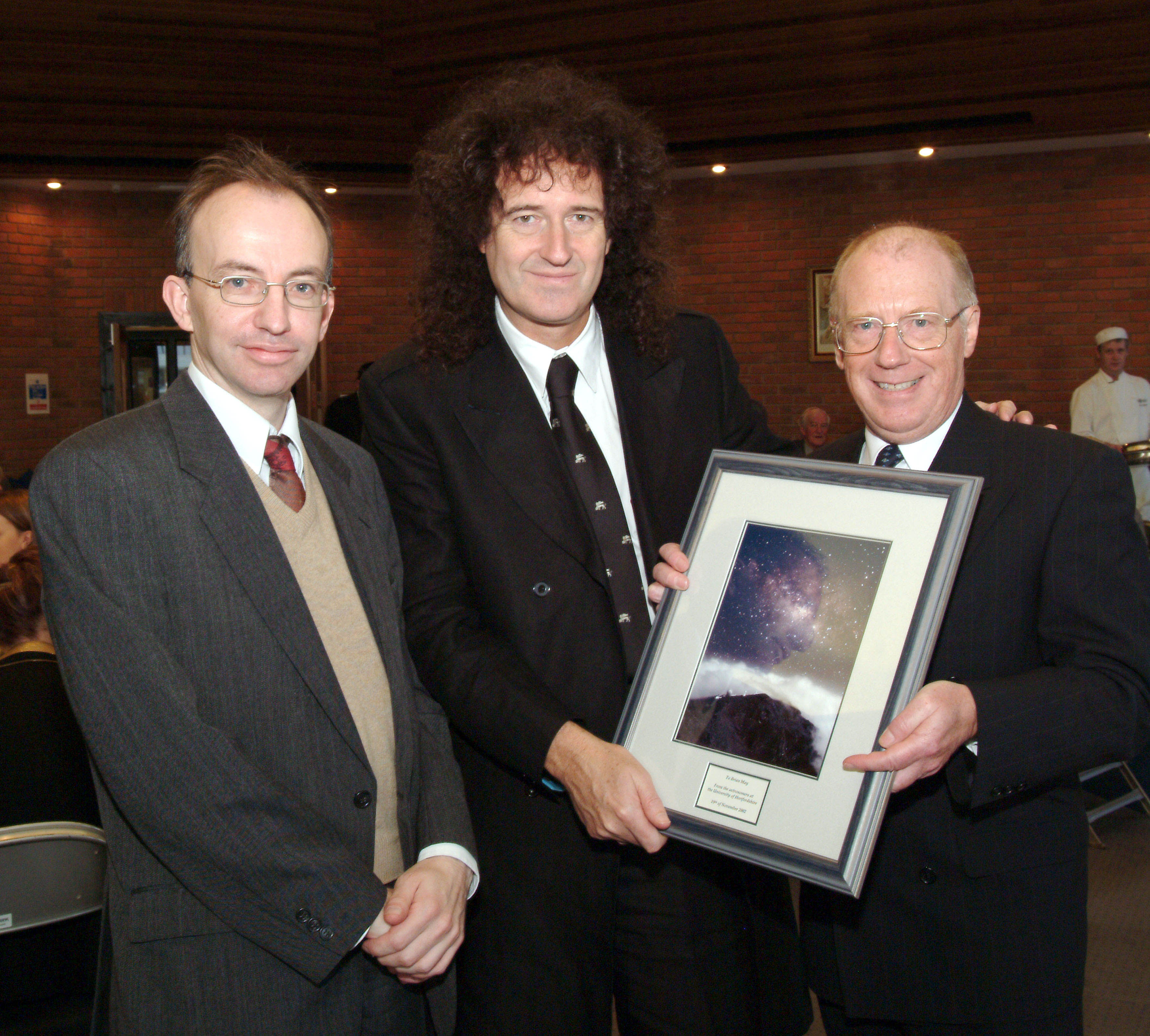 The photo shows Brian May being presented with an astronomical photograph by James Hough and colleague James Collett after the honorary degree ceremony.