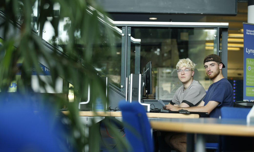 two students looking at the camera whilst sitting at computers in the learning resources centre