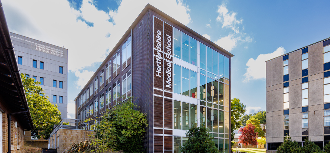 Exterior view of Hertfordshire Medical School at the University of Hertfordshire. The modern four-storey building features extensive glass windows and dark wood cladding, with "Hertfordshire Medical School" displayed vertically on the corner façade. Trees and landscaped greenery surround the building on a bright, sunny day with blue skies and scattered clouds.