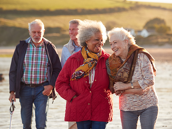 Four older people smile and laugh as they walk along the beach in the sunshine