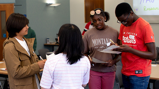 A group of four young people in a huddle are talking whilst one person writes on a piece of A4 papert writing