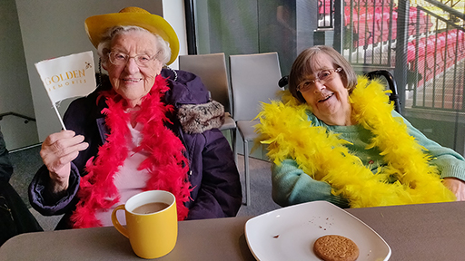 Two mature ladies smile as they enjoy a cuppa and biscuits. One waves a small flag which reads 'Golden Memories'