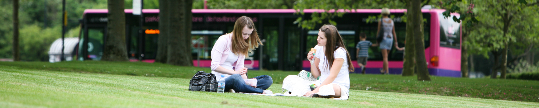 Two young students enjoying a picnic on the lawn. In the distance are trees and a pink Uno bus driving past.