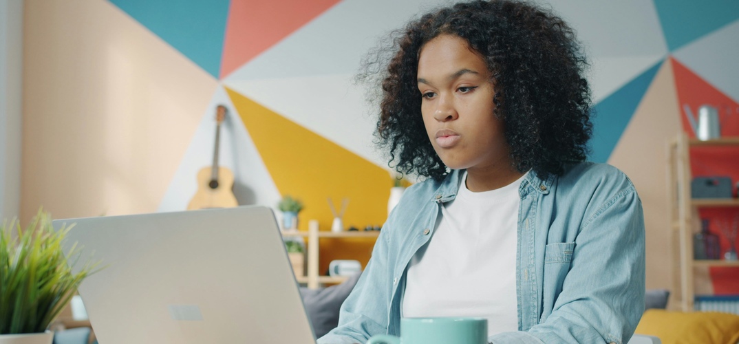 A lady sits on her laptop in a colourful room with a mug and notebook by her side