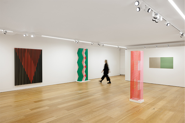 Gallery installation of Rana Begum's minimalist works– a vertical red mesh sculpture in the centre and three wall- based pieces surrounding it as a blurry figure walks through the gallery space.
