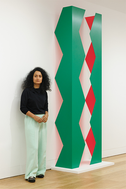 Rana Begum wears a black top and pale green trousers as she stands in front of her vertical, minimalist wall-based green and red sculpture.